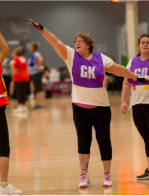 A woman throws a ball towards a netball post while three other women stand with their arms up between her and the Netball post
