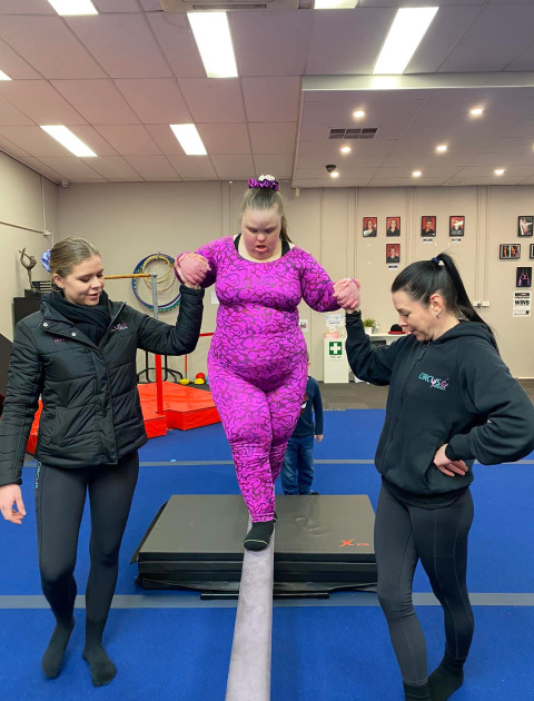 a girl in a pink outfit is walking along a beam, supported on each side by ladies wearing black outfits