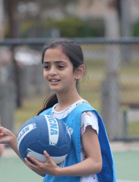 a girl wearing a blue netball bib is smiling at the camera and holding a blue and white netball