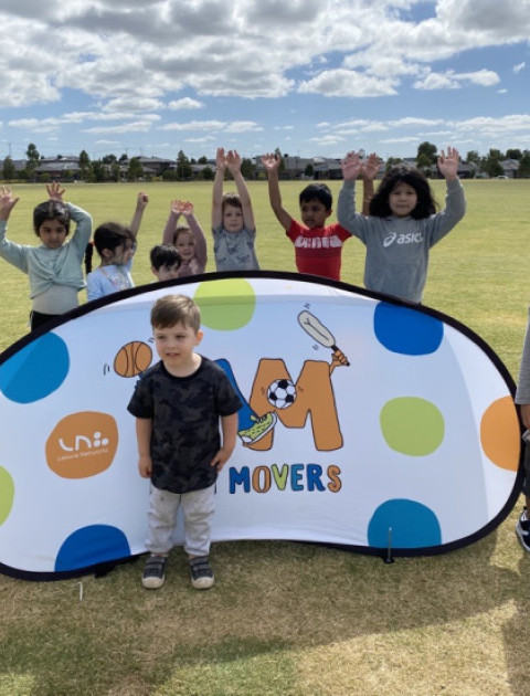 Little kids are standing around a Mini Movers sign holding up their hands