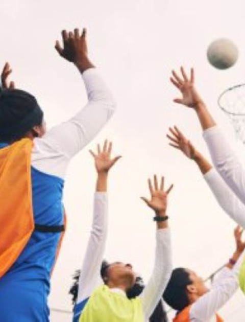 a group of girls wearing netball bibs are reaching up to a netball in the air