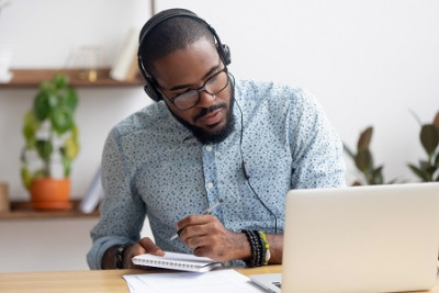 Black man wearing headphones & looking at a laptop takes notes with pen & paper