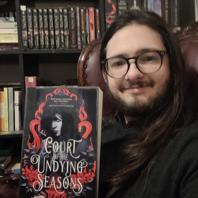 Librarian Steven holds a copy of Court of the Undying Seasons in front of a bookshelf