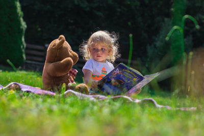 Young girl sitting next to bear reading a book outside