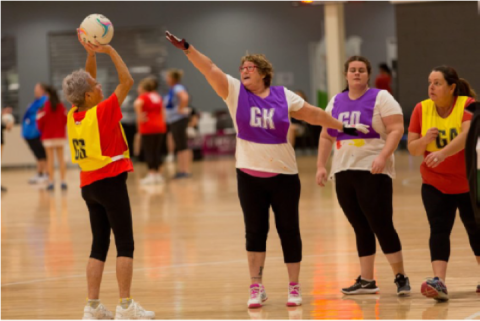 A woman throws a ball towards a netball post while three other women stand with their arms up between her and the Netball post