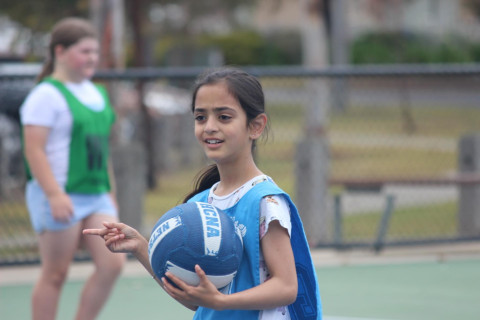 a girl wearing a blue netball bib is smiling at the camera and holding a blue and white netball