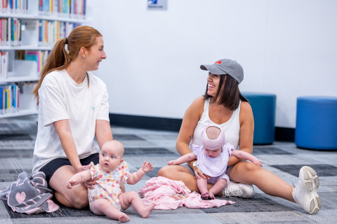 Two mothers and babies sitting in the library and smiling.