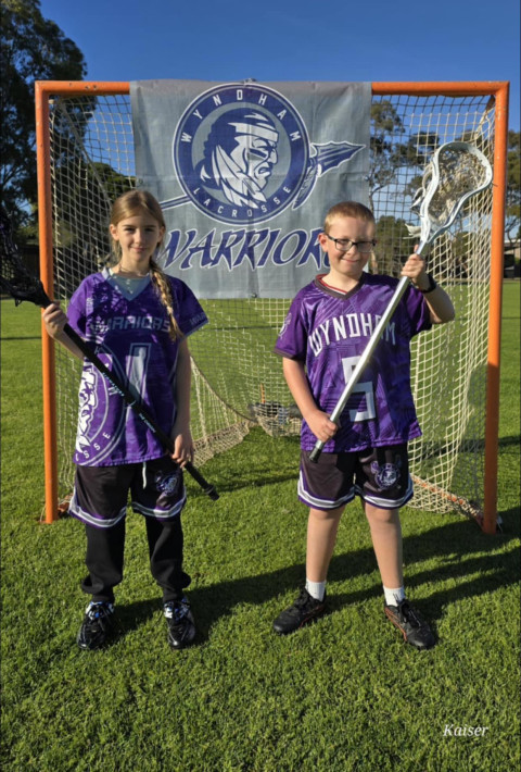 a boy and girl in purple and black sports uniforms stand infront of a goal holding Lacrosse sticks