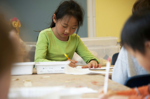 A girl writing on paper in a bright room with other children.
