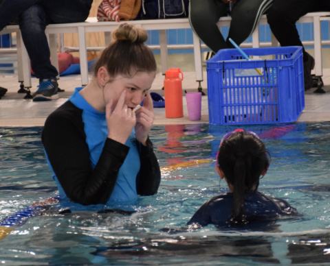 a swimming instructor point to her eyes whilst a child watches, both are in the pool
