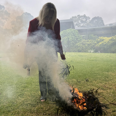 Jillian West conducting Cultural Smoking at an outdoor location. Her gaze is directed towards the fire as she leans forward.