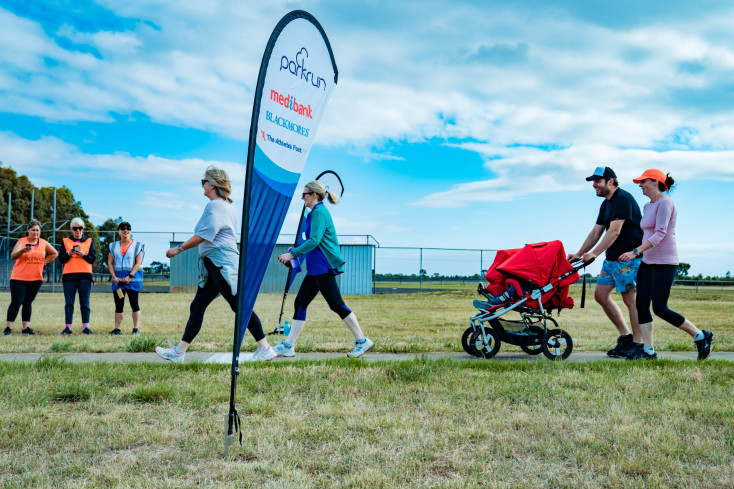 a group of adults standing on grass around a flag which says parkrun are smiling