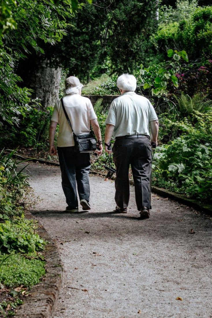 two people with white hair wearing light tops and dark trousers walk along a path away from the camera
