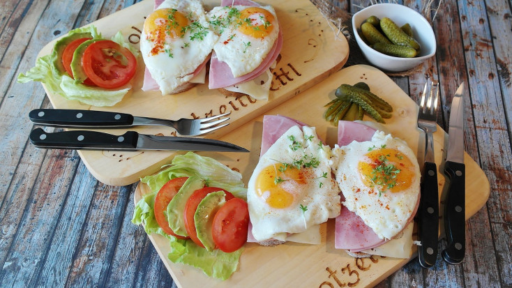 Fried eggs ontop of ham, cheese and bread, with slices of tomato, avocado and lettuce are layed out on wooden bread boards with cutlery on the side