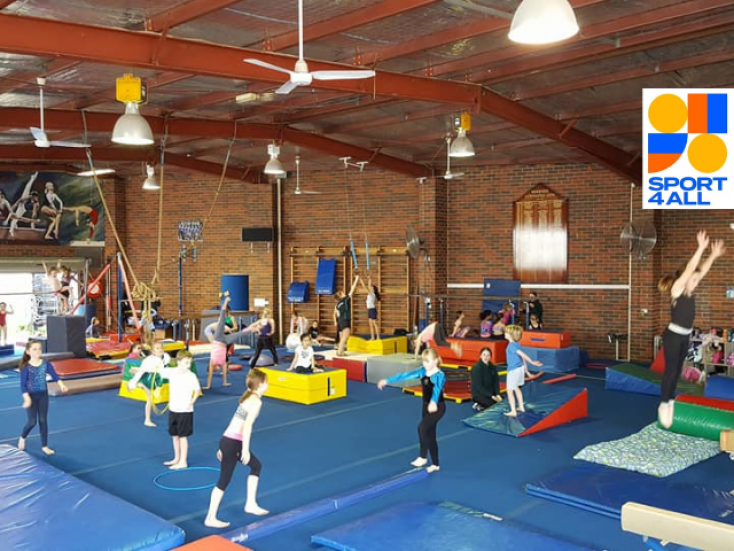 children are spread around different activity stations on blue mats, one is jumping, another has a hoop, another is balancing on a blue beam, another is trying a handstand