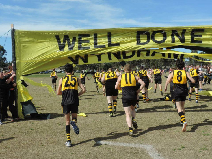 a group of men wearing yellow and black sports clothing run under a yellow banner saying Well Done 