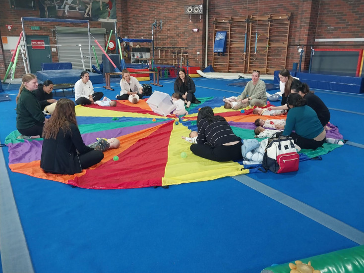 a group of adults sit around a parachute whilst babies crawl ontop