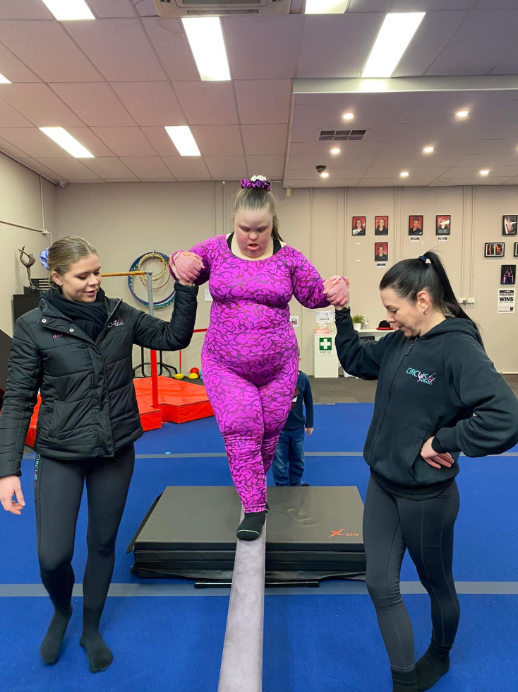 a girl in a pink outfit is walking along a beam, supported on each side by ladies wearing black outfits