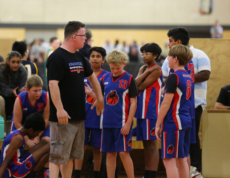 a groups of boys wearing blue and red Basketball uniforms are listening to a coach. One is high fiving the coach.