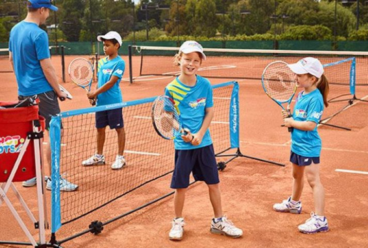 a group ok kids holding tennis rackets stand around a mini tennis net