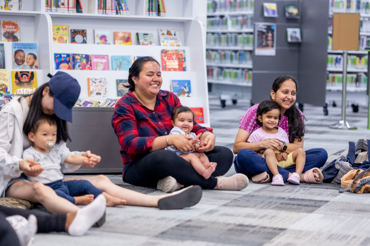 Mums and children sitting on the floor in the library and smiling.