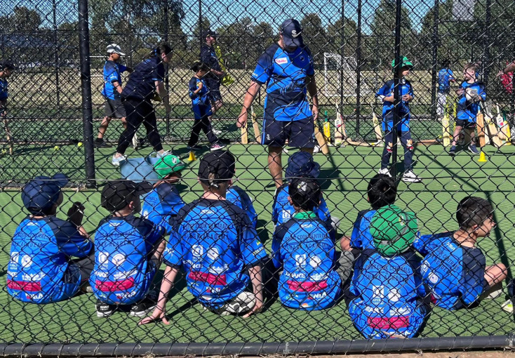 a group of boys are sitting in the cricket nets watching a coach
