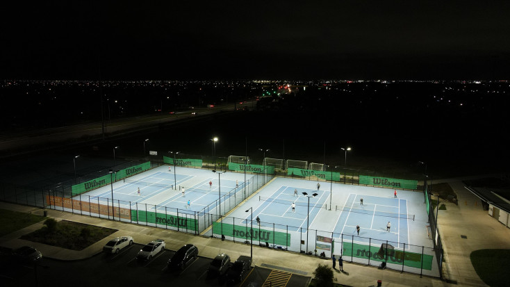 on overhead shot of 4 tennis courts with their lights and people playing Tennis