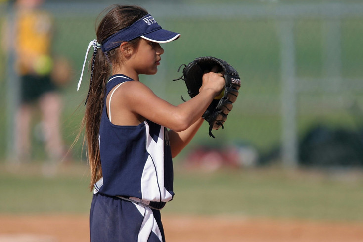 a girl is holdng up a baseball glove and ball, ready to bowl