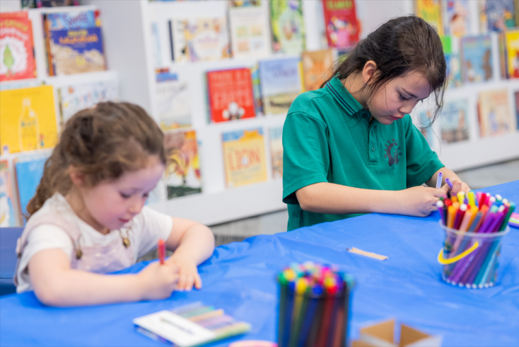 Two girls sit at a table in the library and colour in a craft with bright textas.
