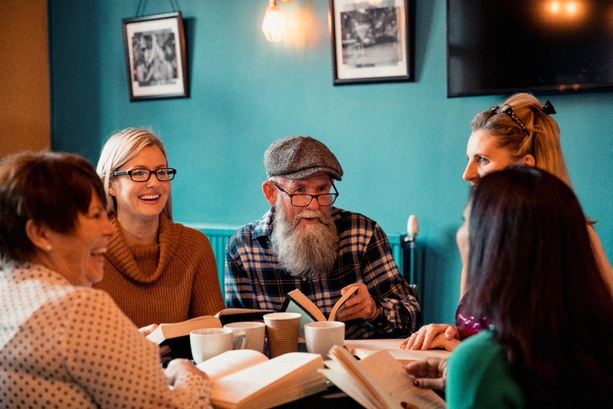 Five people sit around a table with books & hot drinks.