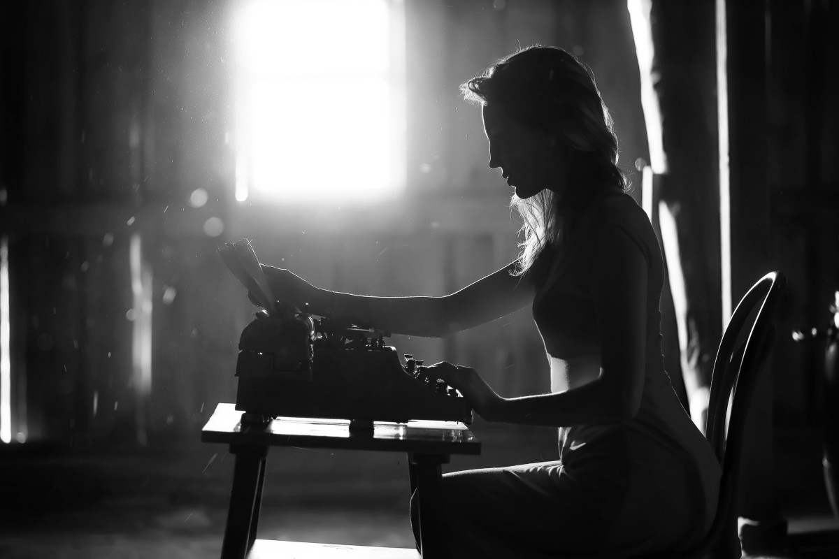 a dramatically lit black and white photograph of a young person sitting at a typewriter