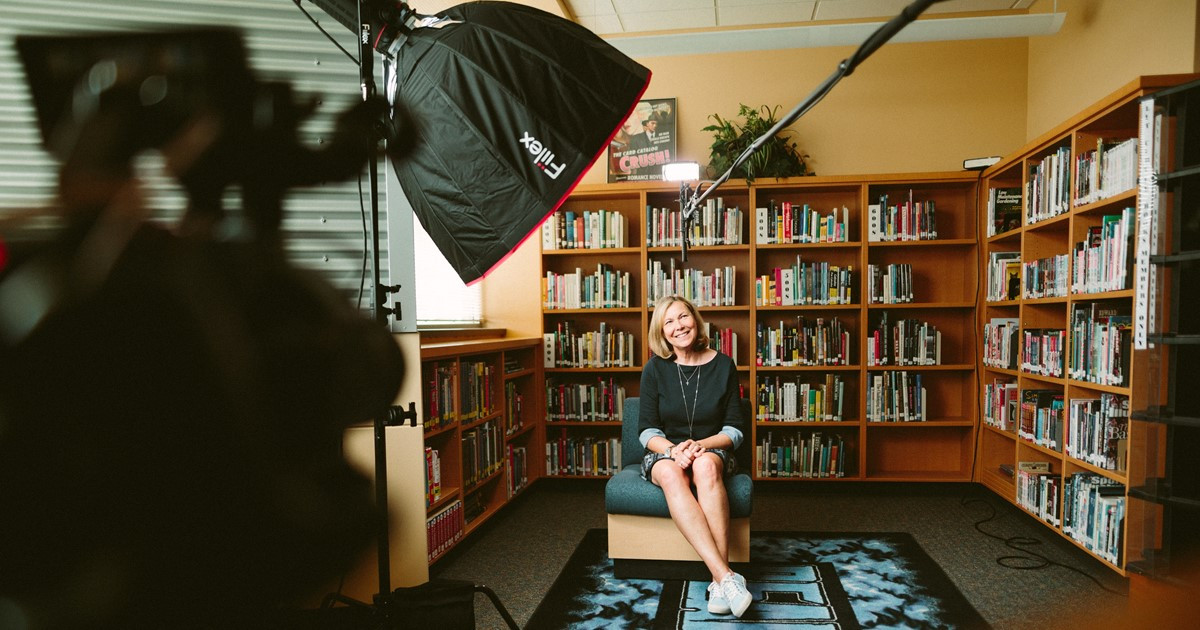 A camera and lighting equipment is ready to record a white woman, seated, in front of bookshelves