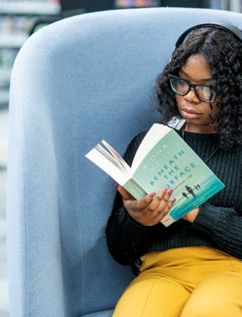 A young person sitting in an armchair, reading, with large headphones on