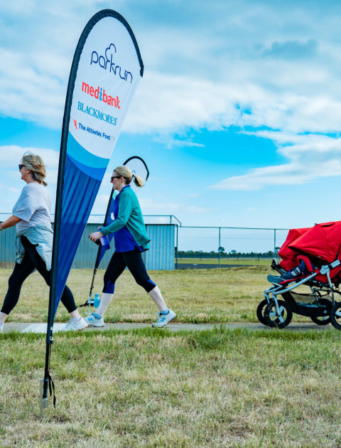 a group of adults standing on grass around a flag which says parkrun are smiling