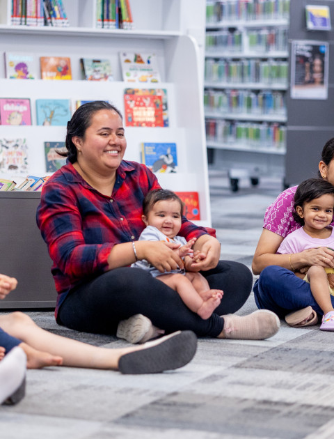 Mums and children sitting on the floor in the library and smiling.