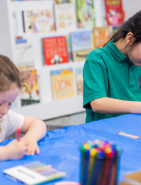 Two girls sit at a table in the library and colour in a craft with bright textas.