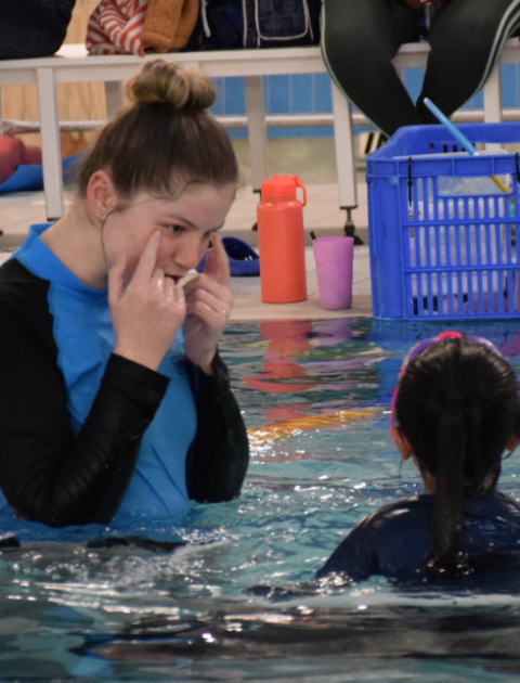 a swimming instructor point to her eyes whilst a child watches, both are in the pool