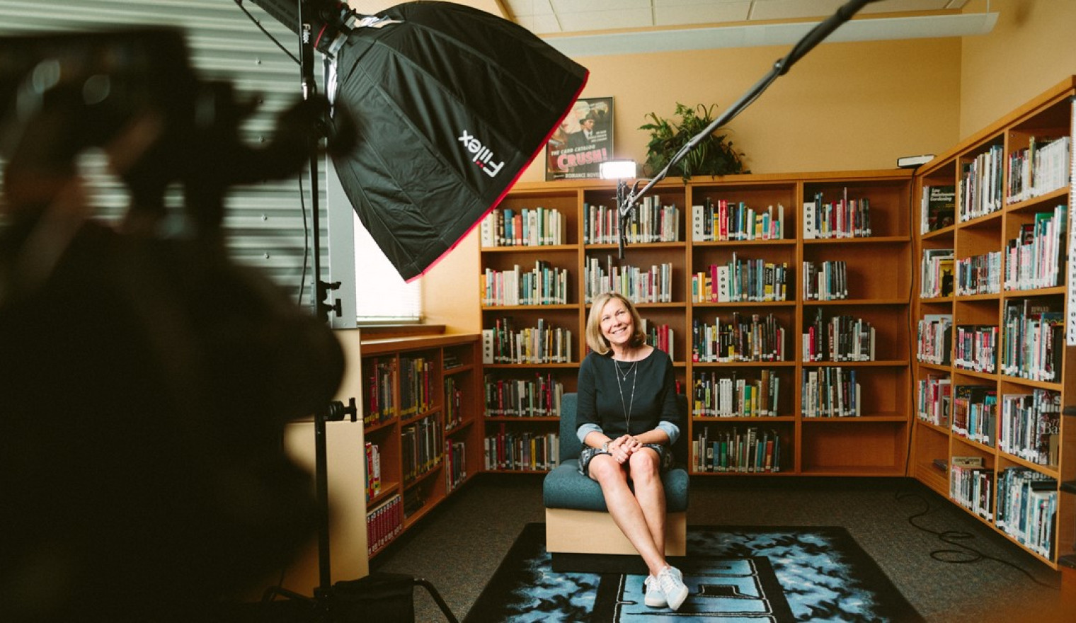 A camera and lighting equipment is ready to record a white woman, seated, in front of bookshelves