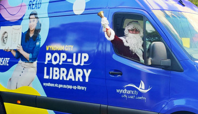 A bright blue Wyndham City Pop-Up Library van with colorful graphics and text promoting reading and creativity. The van features the Wyndham City logo and website address, and a person dressed as Santa Claus is visible through the driver’s window holding a bell.