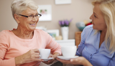 Two adults sitting together indoors, holding cups of tea and talking in a comfortable living room setting with soft furnishings and decor in the background.