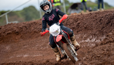 a woman wearing black leather motorcycle clothing and a black helmet is riding a red motorcycle on a dirt track
