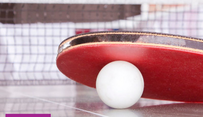 A red table tennis bat rests on a white table tennis ball infront of a table tennis net with This  Girl Can logos at the bottom on a purple background