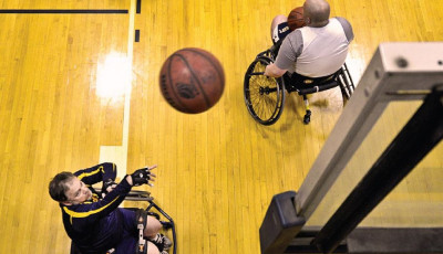 a man in a wheelchair has thrown a basketball up to a basketball hoop, another man in a wheelchair is holding a basketball