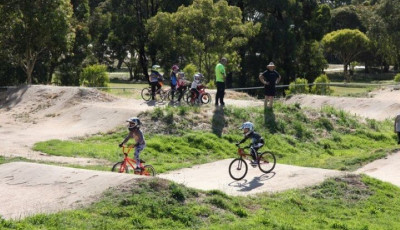a group of children wearing bike helmets and on BMX bikes are ontop of a hill part of the BMX track with 2 adults, two other children are cycling on another part of the BMX track