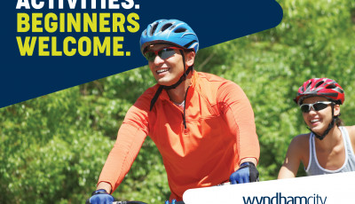A man wearing an orange top and blue cycling helmet is cycling beside a woman wearing a red helmet