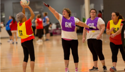 A woman throws a ball towards a netball post while three other women stand with their arms up between her and the Netball post