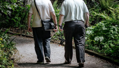 two people with white hair wearing light tops and dark trousers walk along a path away from the camera