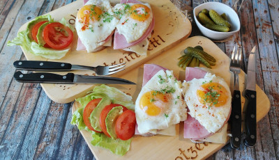 Fried eggs ontop of ham, cheese and bread, with slices of tomato, avocado and lettuce are layed out on wooden bread boards with cutlery on the side