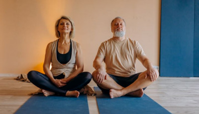 a man and women in pale casual tops and black bottoms site crossed legs on blue mat, side by side with their eyes closed