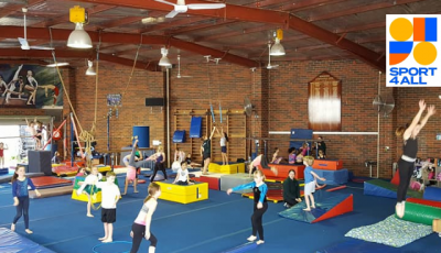 children are spread around different activity stations on blue mats, one is jumping, another has a hoop, another is balancing on a blue beam, another is trying a handstand